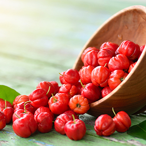 A wooden bowl on its side with bright red Acerola cherries pouring over a wooden tabletop.