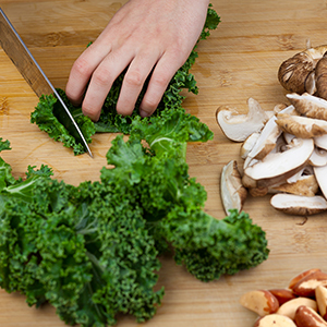 A person slicing kale over a wooden cutting board with mushrooms and Brazil nuts.